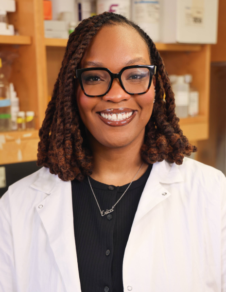 Woman with brown hair wearing a white coat and black glasses smiling in a lab setting